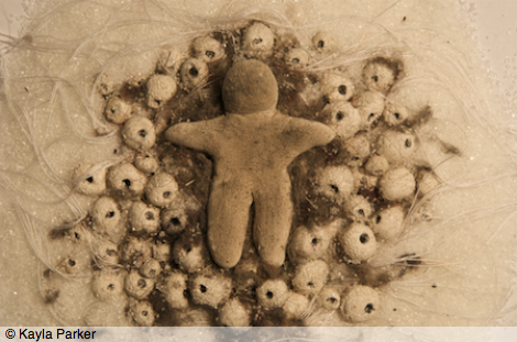 film frame from White Body depicts close-up image of small figure or doll (no features) sculpted from white modeling clay, lying in a bed of white crystals (granulated sugar), white sewing thread, and tiny round white eggs/beads, infiltrated with grey-brown dusty fluff (slut's wool)