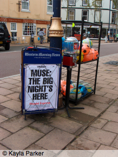 Photo of triangular Western Morning News 'headlines' sign for local newspaper the Herald Express, on pavement by lamppost outside shop; sign reads: