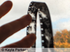 Close-up colour photo of Kayla Parker's left hand on the window in Studio One, with loop of 16mm film; sky and trees behind