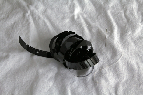 Colour photograph is of a length of black 16mm film, single perf, exposed to no light and processed, unraveling from the core, with a sewing needle threaded with white cotton; against a background of a white cotton sheet.